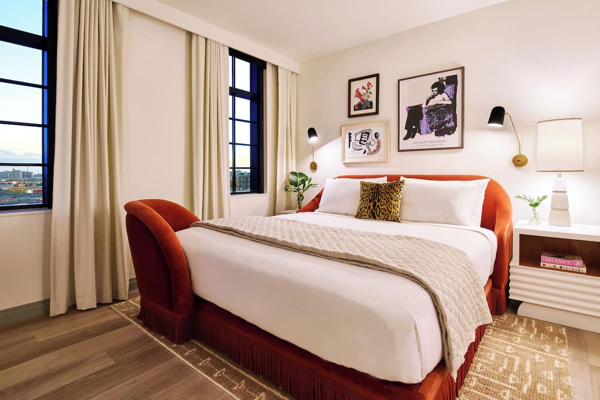 Une chambre d’hôtel avec un grand lit, un fauteuil rouge, une table de chevet avec une lampe et deux œuvres d’art encadrées au mur.
