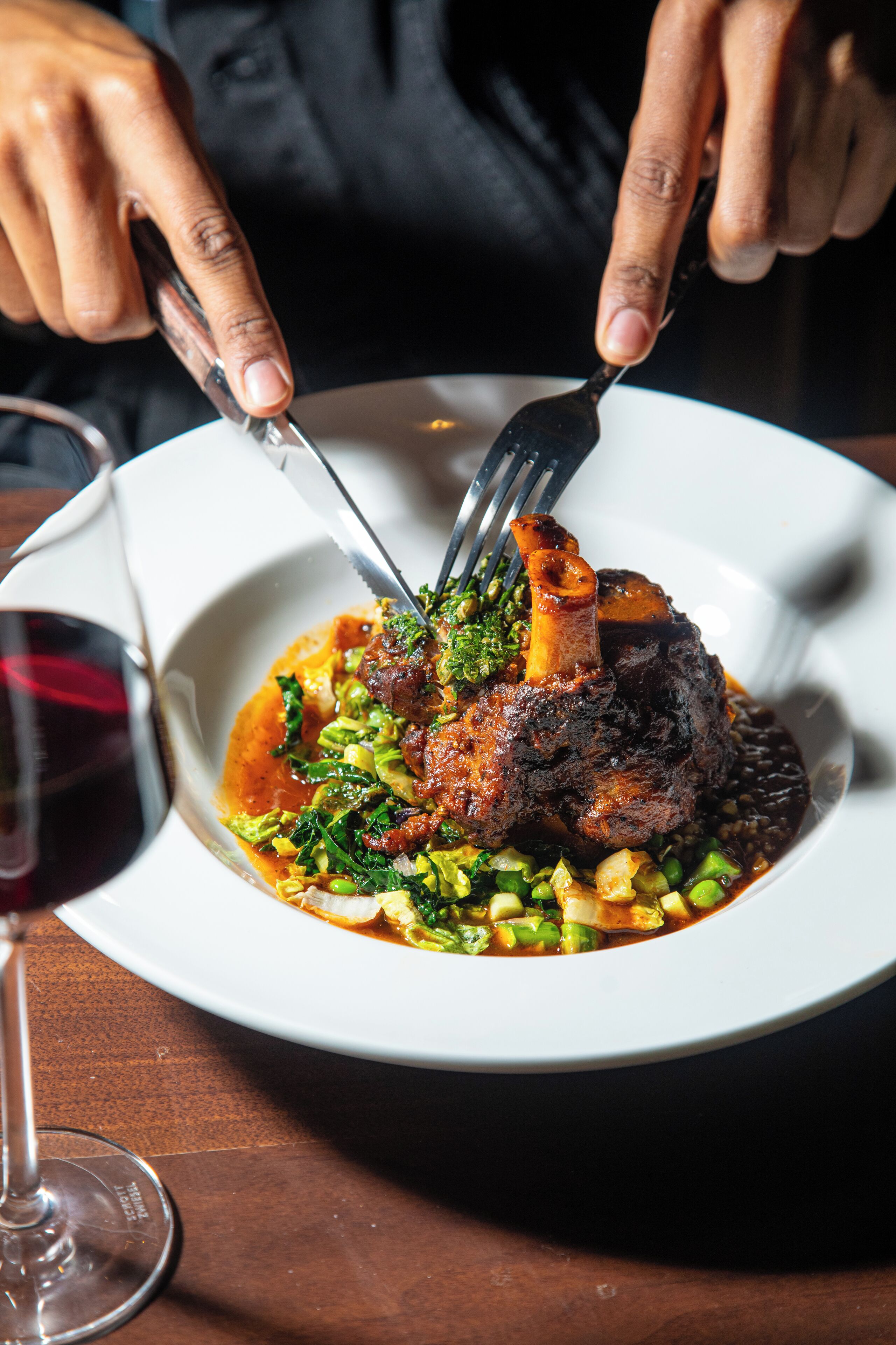 A person dining with a fork and knife on a plate of food, a glass of red wine, and a wooden table.