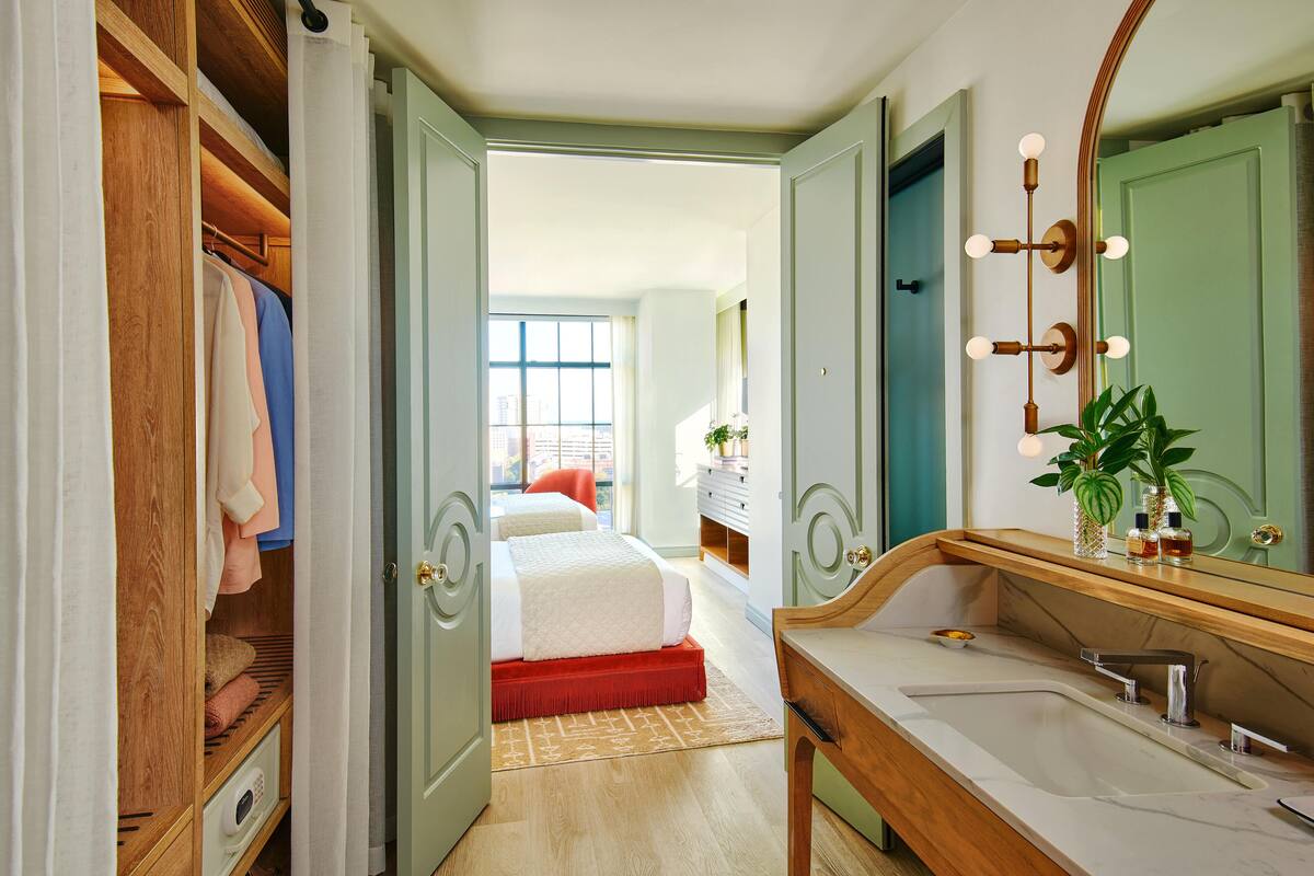 Une chambre avec une armoire en bois, un lit agrémenté d’un couvre-lit rouge, une salle de bain avec un miroir, un lavabo et une plante.