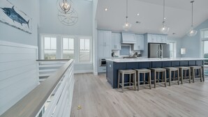 A spacious kitchen with a large island, light blue cabinets, and a wooden railing overlooking a balcony. - 5848- Sun & Sea time - Oceanfront Escape | Pool, Hot Tub, Sleeps 18 (Nags Head)