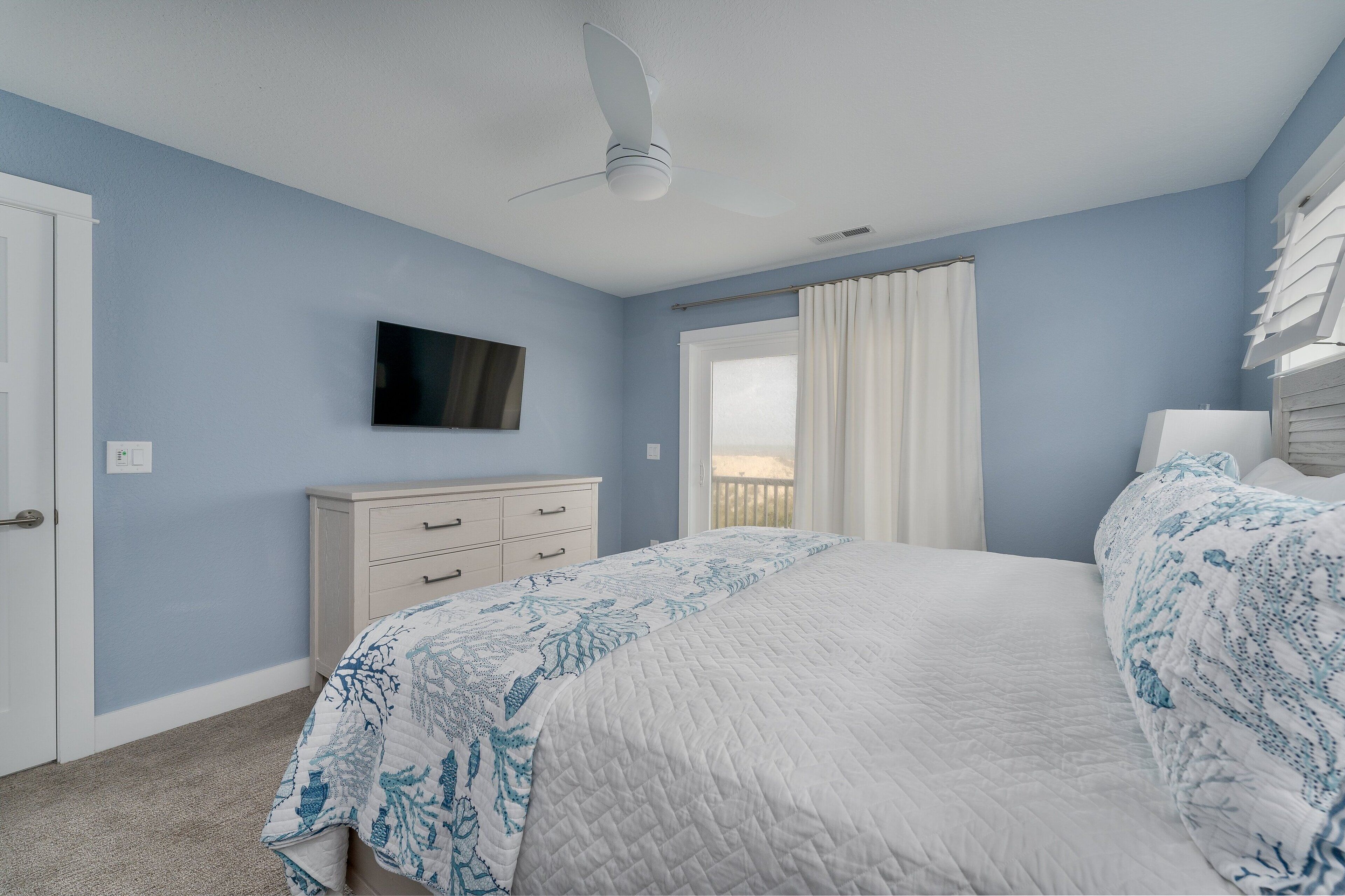 A bedroom with a bed, a ceiling fan, a window with curtains, a dresser, and a television on the wall.
