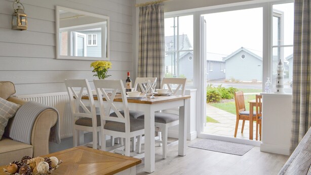 A dining area with a table set for four, white chairs, a wooden table, and a view of the outside through a large window.