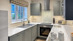 A modern kitchen with grey cabinets, a built-in oven, a stainless steel range hood, a double sink, and a window with a view of greenery.