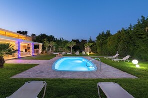 A swimming pool with lounge chairs, a stone deck, and a modern house with illuminated columns.