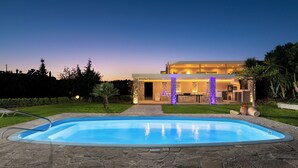 A swimming pool with stone tiles, a patio, and a modern house with a well-lit entrance.