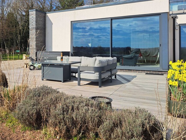 A modern outdoor living area with a sofa, coffee table, and chairs, a large sliding glass door, and a wooden deck.