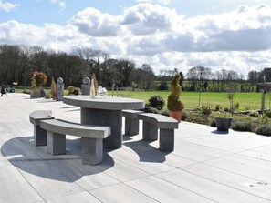 A stone outdoor seating area with benches and planters, set against a backdrop of a green field and trees. - A stylish converted barn annex. (Lichfield)