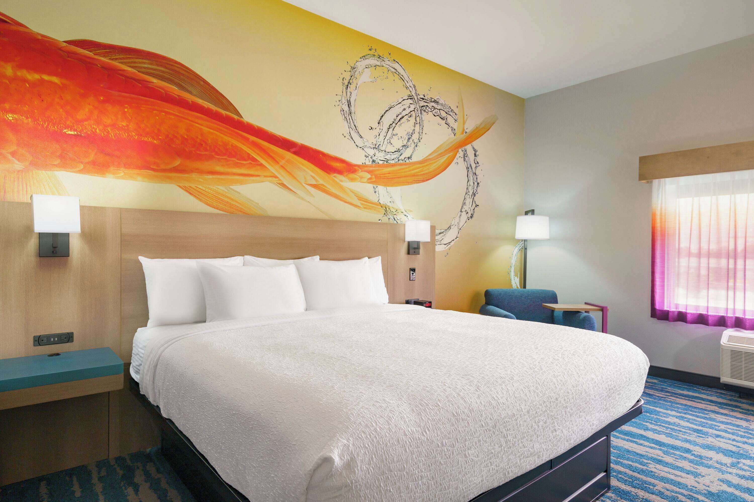 Une chambre d’hôtel avec un grand lit, une tête de lit, une table de chevet, une lampe, une chaise et une fenêtre avec des rideaux.