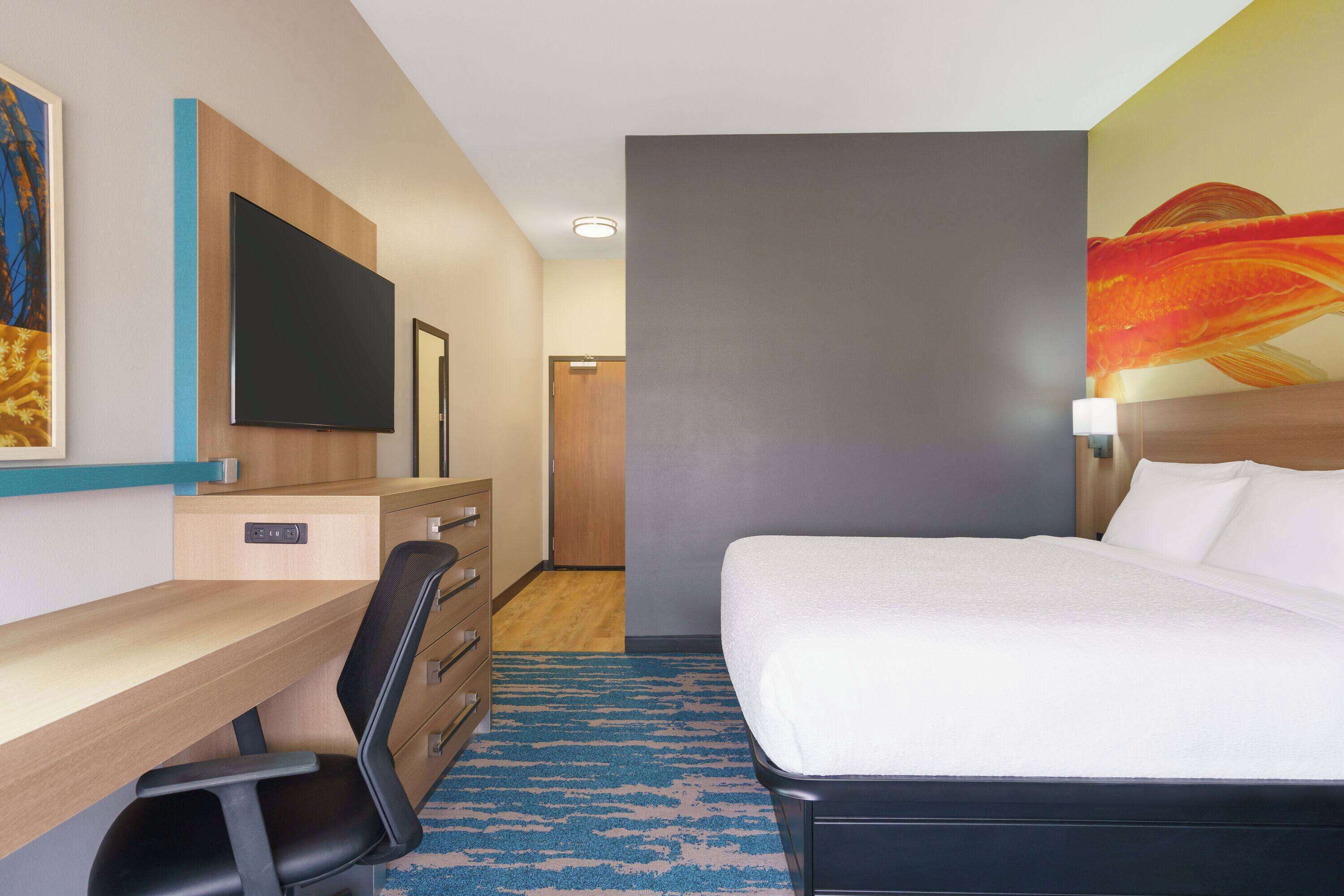 Quarto de hotel com cama, escrivaninha, cadeira, televisão e obras de arte nas paredes.