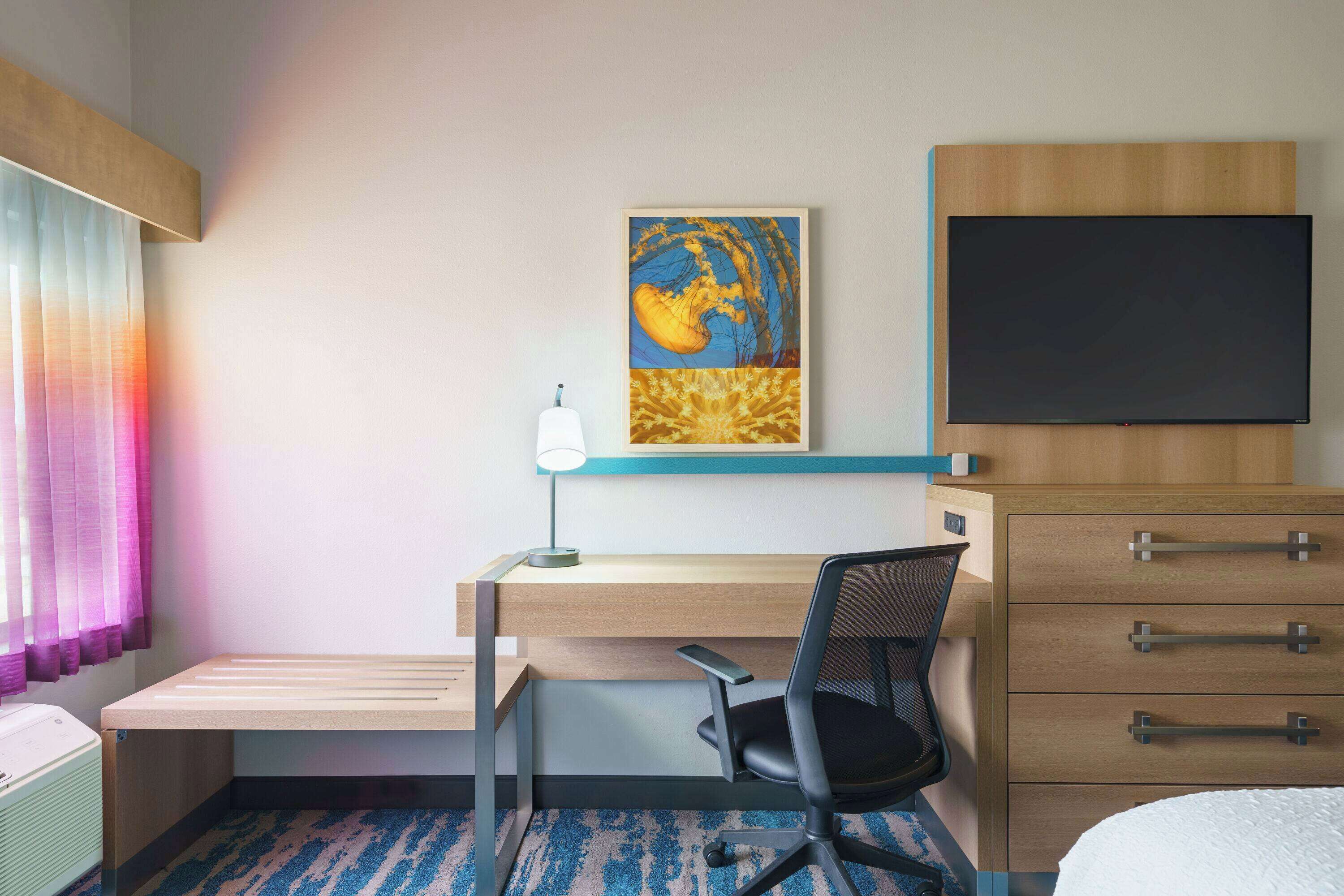 Une chambre d’hôtel comprenant un lit, un bureau avec une chaise, une télévision, un tableau et une commode.