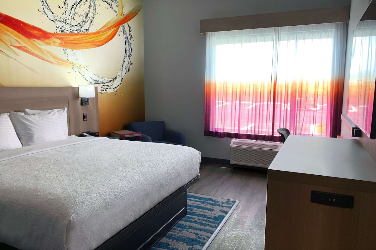 Una camera d'albergo con un letto, una scrivania, una sedia, una finestra con tende e un quadro appeso al muro.