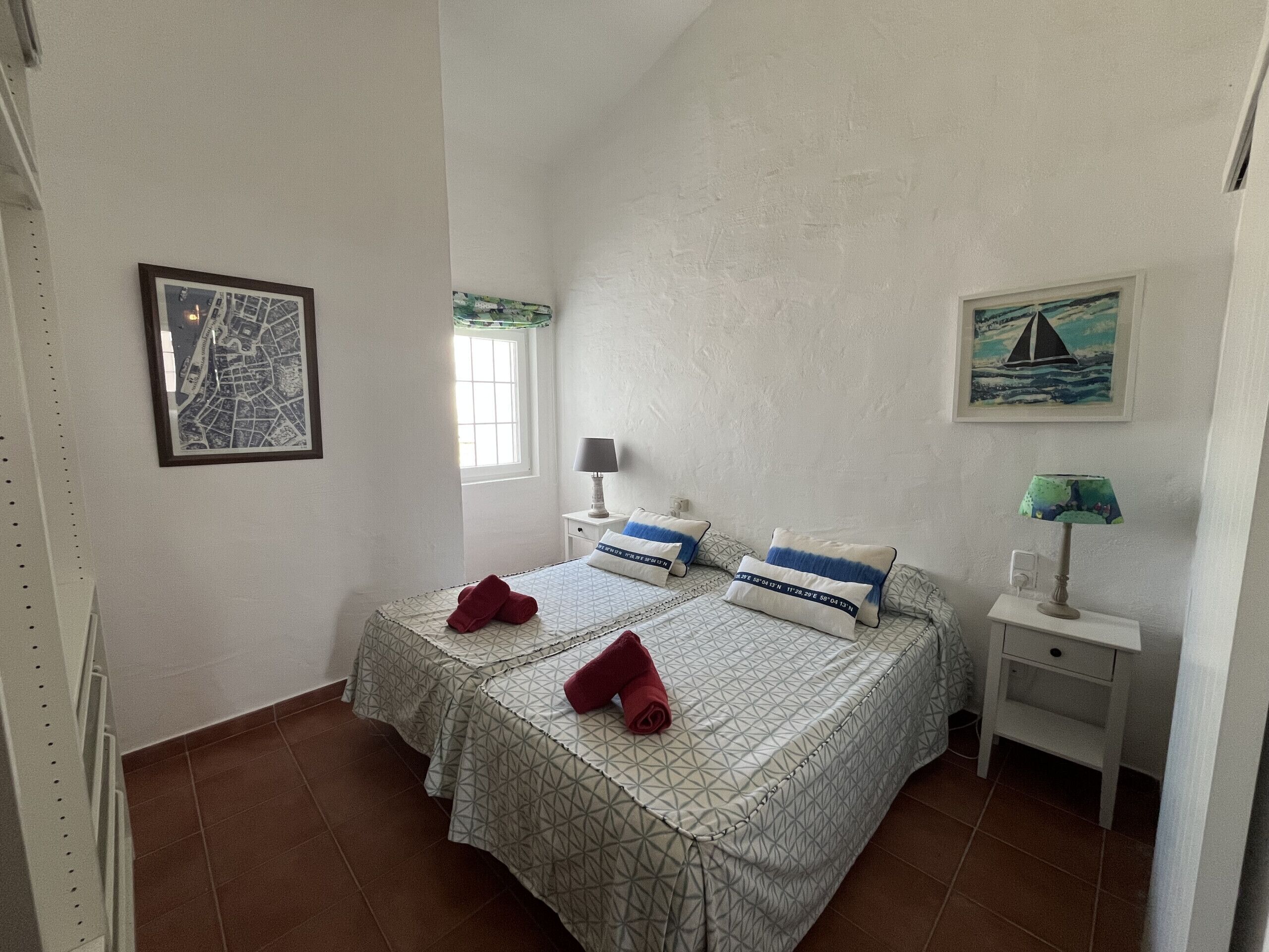 A bedroom with two beds, a window, a nightstand, a lamp, and two wall-mounted paintings.