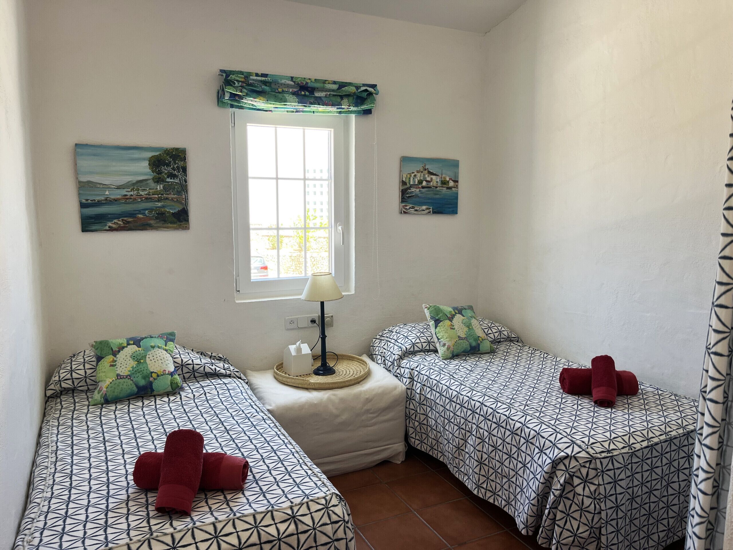 A room with two beds, each with patterned bedding and pillows, a small table with a lamp, and two paintings on the walls.