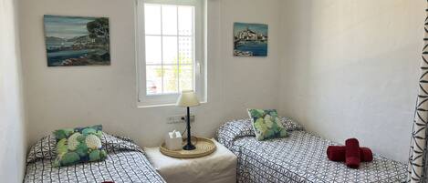 A room with two beds, each with patterned bedding and pillows, a small table with a lamp, and two paintings on the walls.