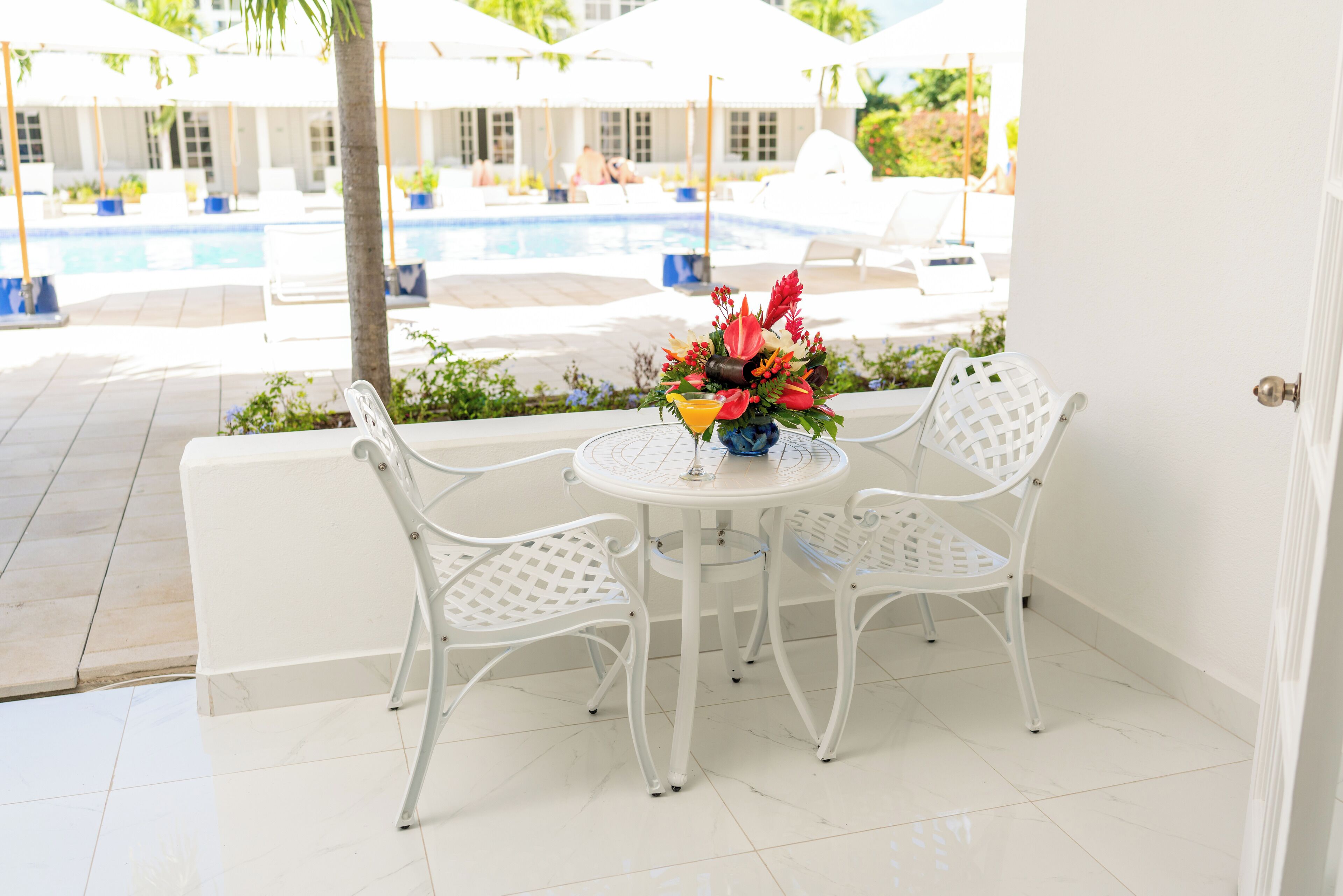 A white outdoor seating area with a table and chairs, a flower arrangement, and a view of a pool and lounge chairs in the background.