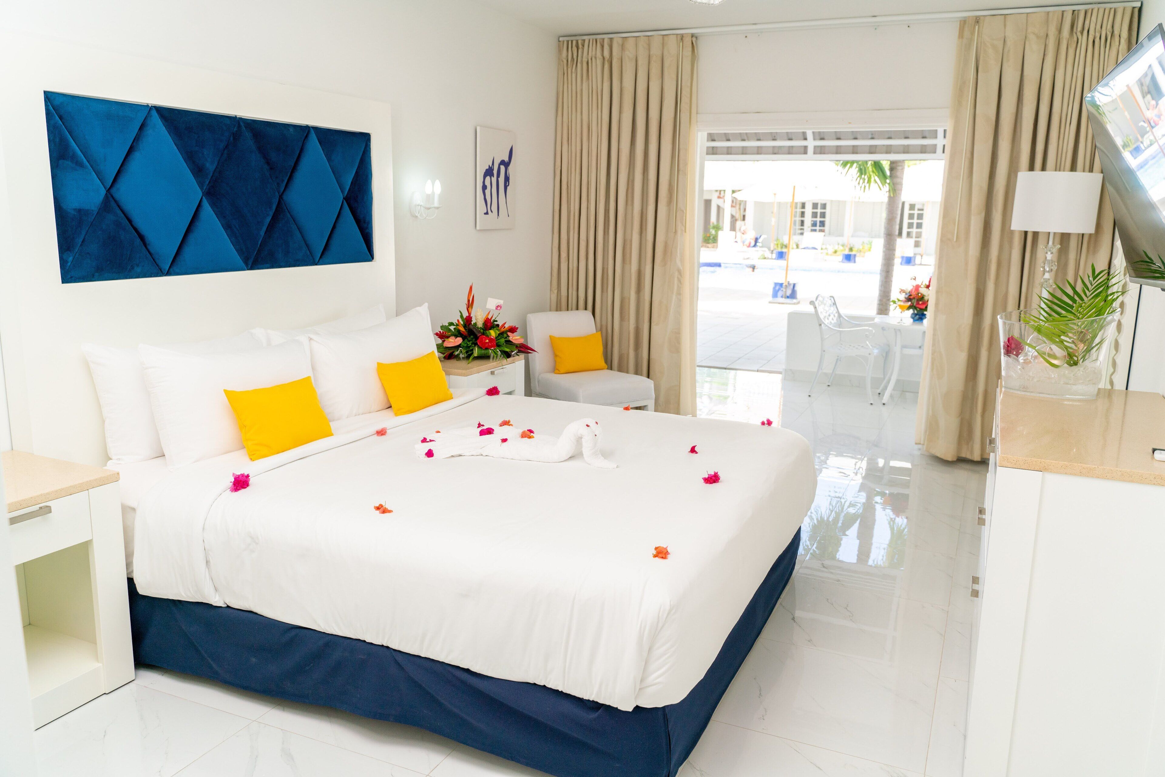 A hotel room with a bed, white and yellow pillows, a blue accent wall, a flower arrangement, and a view of the outside.