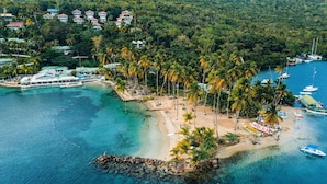 An aerial view of a tropical island with a beach, palm trees, boats, and a resort area with buildings.