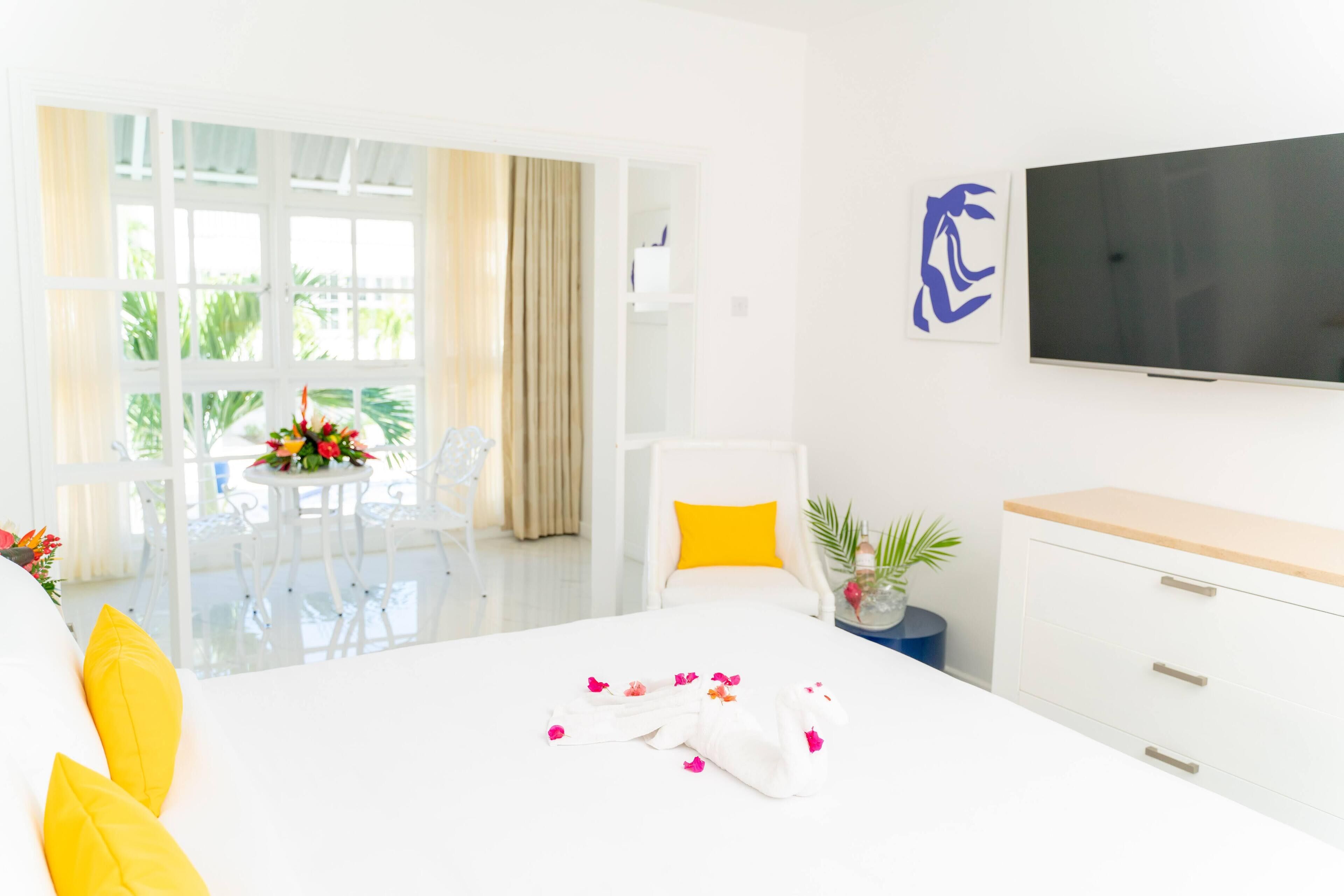A hotel room with a bed, a TV, a desk, and a chair. There is a window with curtains, a plant, and a table with flowers.