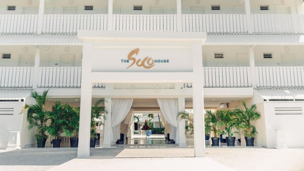 A hotel entrance with a sign reading "The Suc House," featuring a covered walkway, potted plants, and a clear sky.