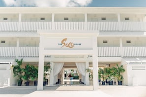 A hotel entrance with a sign reading "The Suc House," featuring a covered walkway, potted plants, and a clear sky.