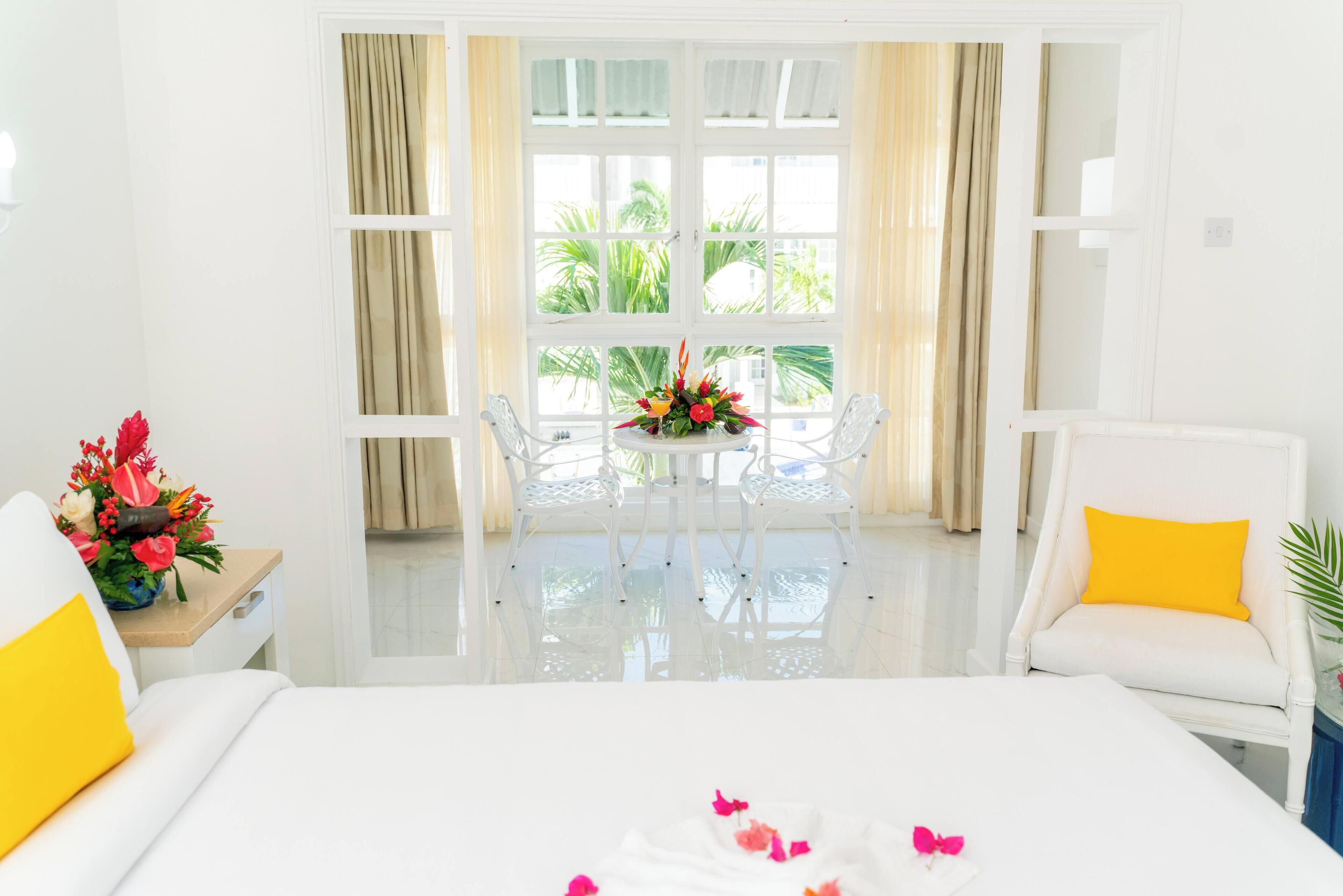 Habitación de hotel con cama, una silla blanca, una mesa con un arreglo floral y vistas a palmeras a través de amplias ventanas.