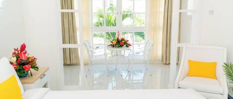 Habitación de hotel con cama, una silla blanca, una mesa con un arreglo floral y vistas a palmeras a través de amplias ventanas.