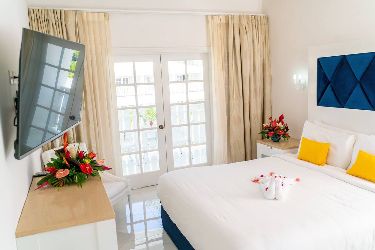 A hotel room with a bed, a TV mounted on the wall, a desk with a flower arrangement, and a view through the door.