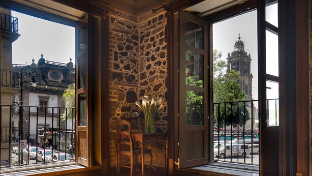 Una habitación con pared de piedra, una mesa de madera con un florero y vistas a una ciudad que incluye una catedral.