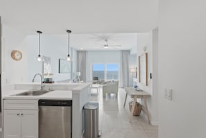Une cuisine moderne équipée d’un lave-vaisselle, un coin repas avec une table et des chaises, et une vue sur la mer grâce à de grandes fenêtres.