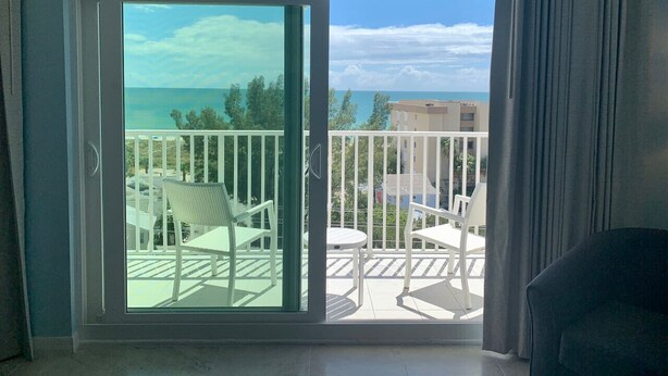 Une pièce dotée d’une porte-fenêtre coulissante donnant sur un balcon agrémenté de meubles blancs et offrant une vue sur la mer.