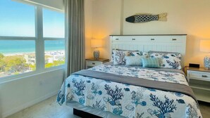 Une chambre avec une grande fenêtre, un lit recouvert d’une courtepointe aux motifs bleus et blancs évoquant l’océan, et un élément décoratif mural placé au-dessus du lit.