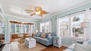 Sala de estar con sofá, mesa de centro y ventilador de techo. Hay ventanas con persianas y vistas al exterior.