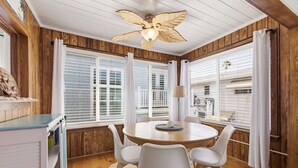 Una habitación con techo de madera, una mesa redonda con sillas, un ventilador de techo y ventanas amplias con persianas.