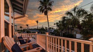 Una terraza de madera con sillas, barandilla y vistas a palmeras y casas bajo un cielo al atardecer.