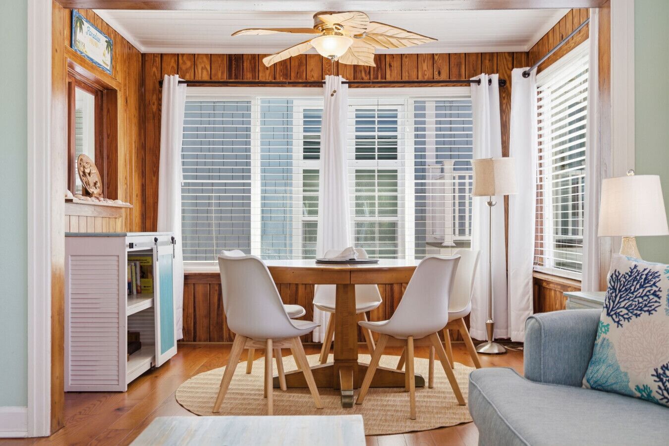 A dining area with a wooden table, four chairs, a ceiling fan, and large windows with shutters.