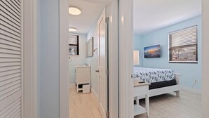 Ein Schlafzimmer mit Bett, Nachttisch, Fenster mit Jalousien, einem an der Wand hängenden Bild und einer Tür zu einem Badezimmer mit Waschbecken und Toilette.