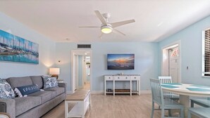 Ein Wohnzimmer mit grauem Sofa, einem Couchtisch, einem Esstisch mit Stühlen, einem Deckenventilator und Kunstwerken an den Wänden.