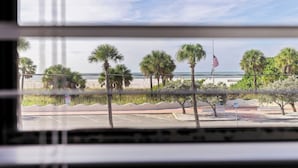 A view from inside a room with a window overlooking a beach with palm trees and an American flag. - Beach-Goers Paradise Getaway! Pool! Sun! Fun! (St. Pete Beach)