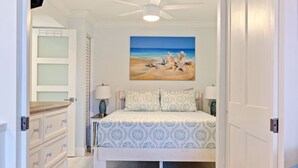 A bedroom with a bed, a nightstand, a lamp, a dresser, and a painting of a beach scene. - Poolside Paradise with Patio, Steps from Beach (St. Pete Beach)