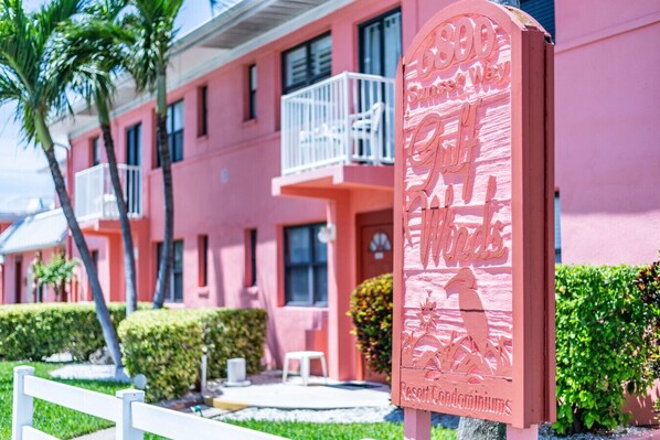 A pink building with balconies, a sign for Sunset Waves Resort Condominiums, and a white fence.