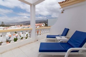 A balcony with two lounge chairs and a small table, overlooking a coastal town with white buildings and a clear sky.