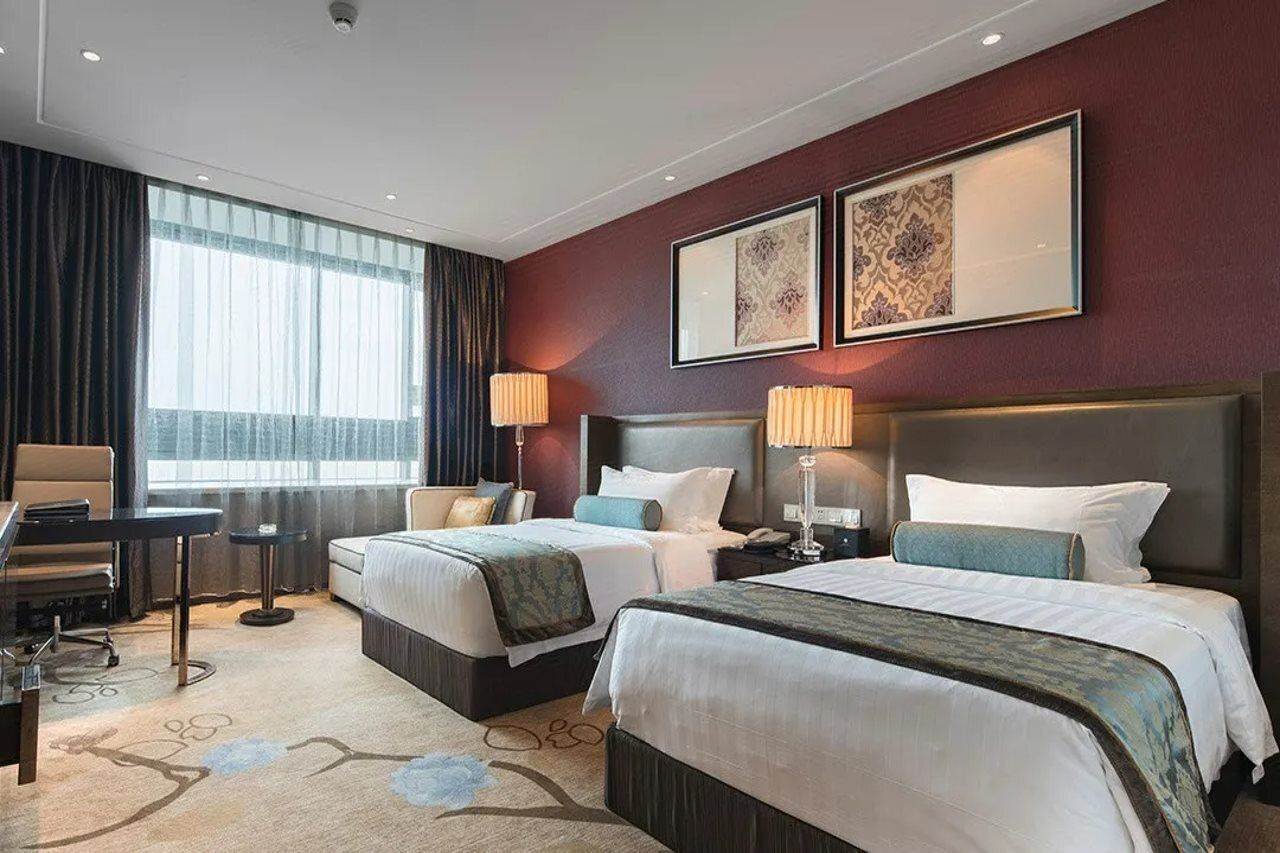A hotel room with two beds, a desk, and a chair. There are two lamps, a window with curtains, and framed artwork on the wall.