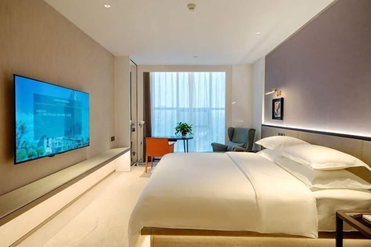 A modern hotel room with a large bed, a flat-screen TV mounted on the wall, a small table with a chair, and a window with sheer curtains.