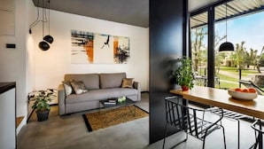 Un salon moderne avec un canapé gris, une table basse et un coin repas comprenant une table et des chaises en bois. Deux tableaux ornent le mur, et une grande fenêtre offre une vue sur l’extérieur.