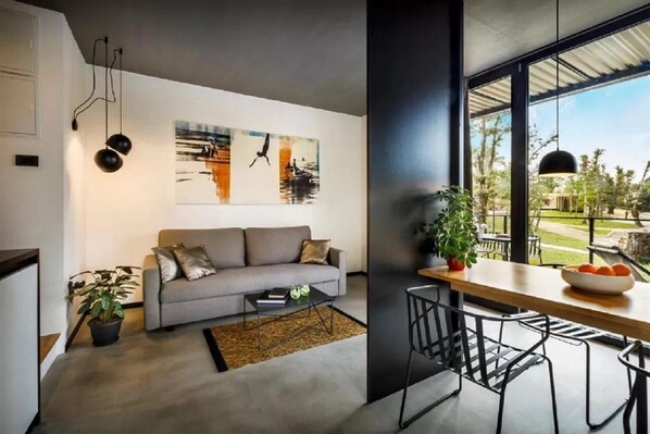 Ein modernes Wohnzimmer mit grauem Sofa, einem Couchtisch und einem Essbereich mit Holztisch und Stühlen. An der Wand hängen zwei Bilder und ein großes Fenster mit Blick ins Freie.
