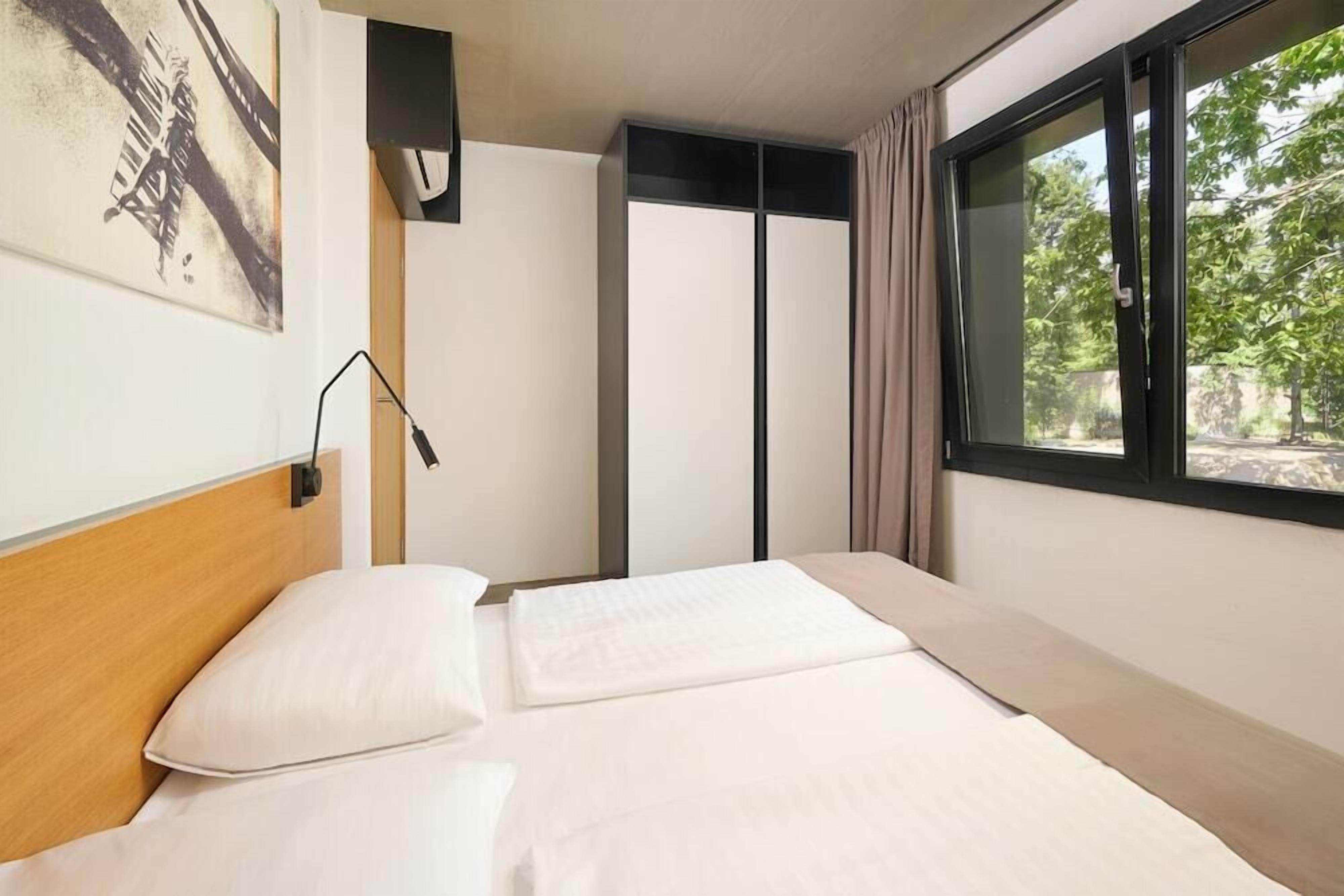 Et hotelværelse med en seng, et natbord, en lampe, et vindue med gardiner og et billede på væggen.