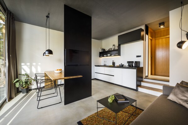 Una cocina moderna con un refrigerador negro, gabinetes blancos y una encimera de madera. Hay una zona de comedor con una mesa y sillas, y un sofá en la sala de estar.