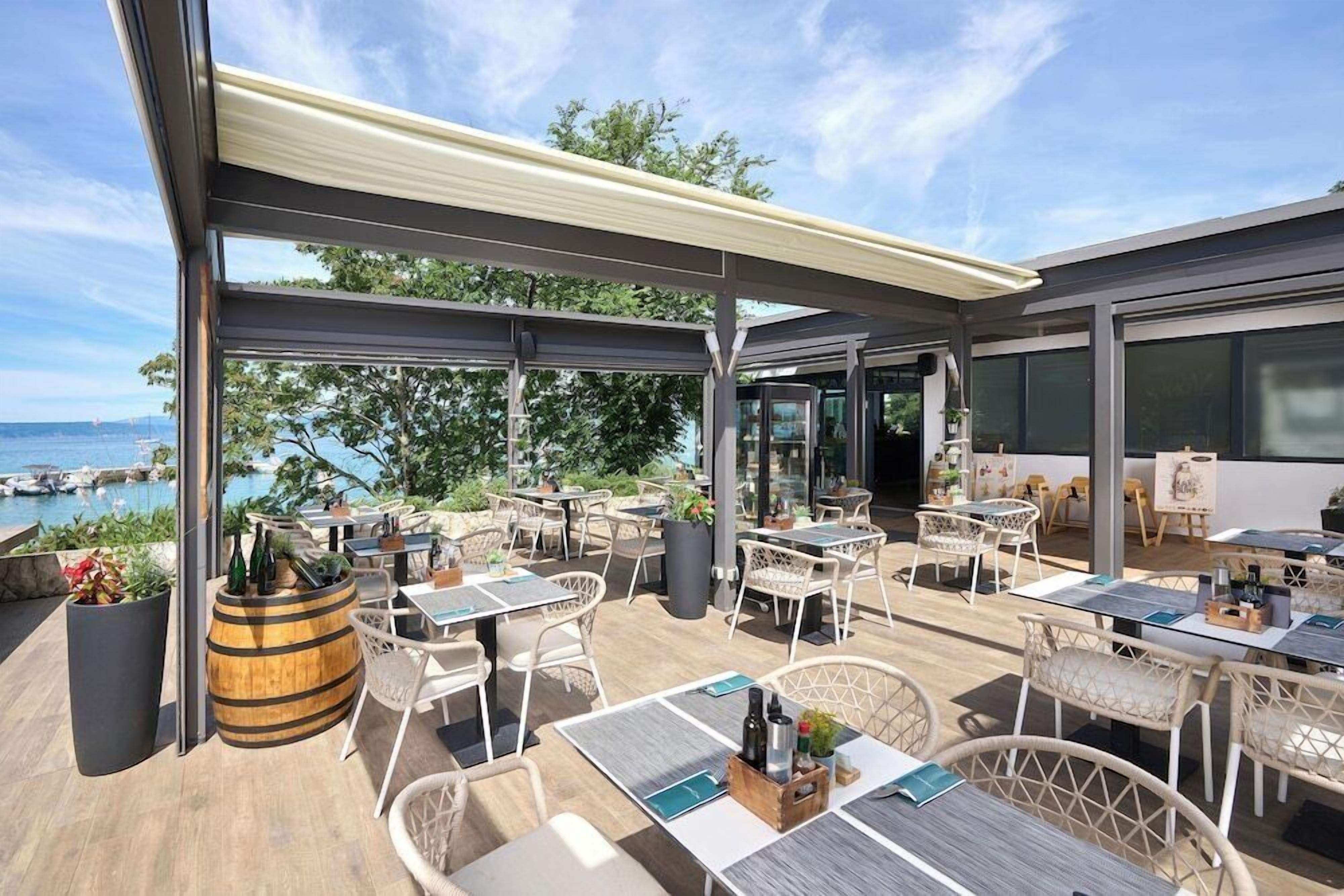 Een terras buiten met tafels en stoelen, een houten vat en uitzicht op de zee.