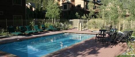 A swimming pool with lounge chairs and a table, surrounded by a brick patio and a wooden fence.