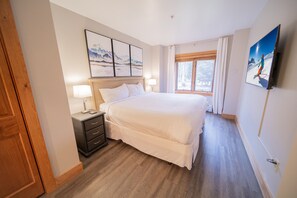 A hotel room with a large bed, a nightstand, a lamp, a window with curtains, and a painting on the wall. - Red Hawk Lodge 2211 (Keystone)